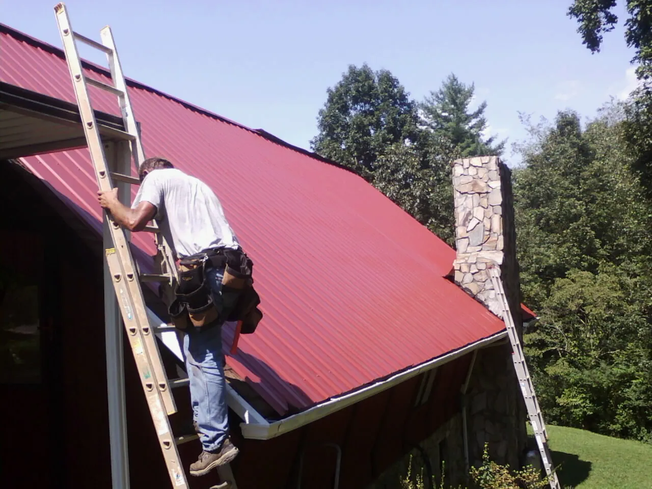 Roofer on a ladder beside a red metal roof and stone chimney — Soffit & Fascia Repair by Roofing Brothers in Gauley Bridge